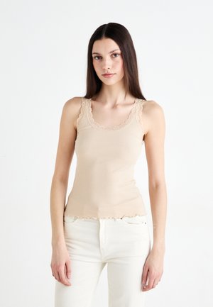 Beige ribbed tank top with lace trim along the neckline and straps, featuring a scalloped hem and fitted silhouette. Paired with white jeans.