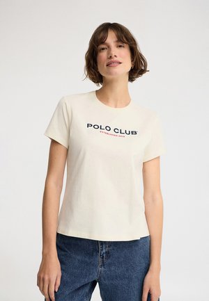 Femme aux cheveux bruns courts portant un T-shirt blanc "Polo Club" et un jean bleu, debout devant un fond blanc uni.