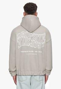 Lys beige hoodie med et stort hvidt trykt logo på ryggen, forsynet med en justerbar hætte og ribbet kant og manchetter.