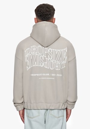 Light beige hoodie with a large white printed logo on the back, featuring a drawstring hood and ribbed hem and cuffs.