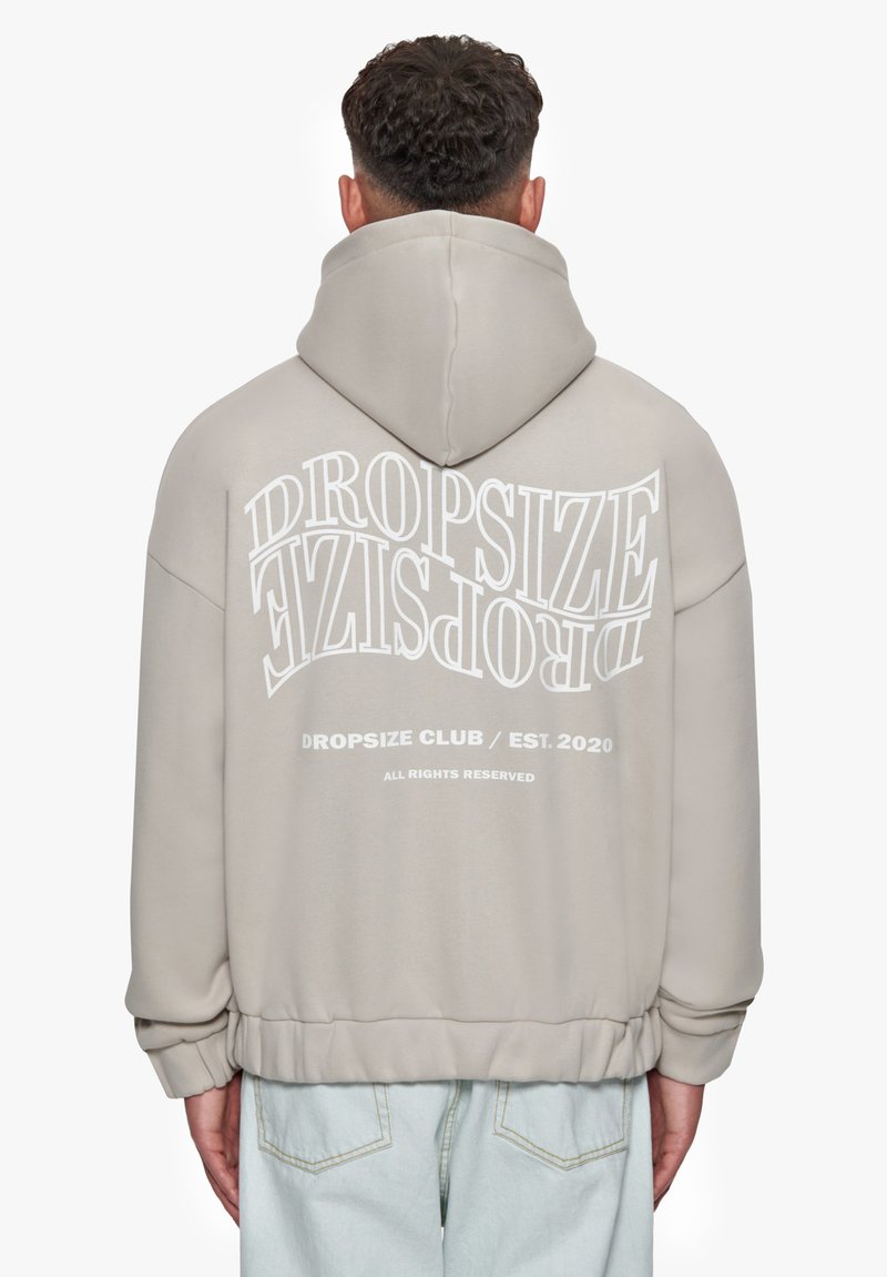 Lys beige hoodie med et stort hvidt trykt logo på ryggen, forsynet med en justerbar hætte og ribbet kant og manchetter.