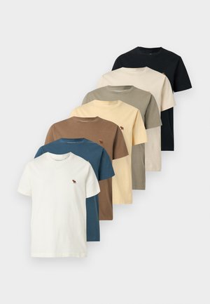 Jeune homme debout avec une main dans la poche, portant une chemise crème à manches courtes avec un petit logo et un jean bleu clair délavé sur fond blanc.