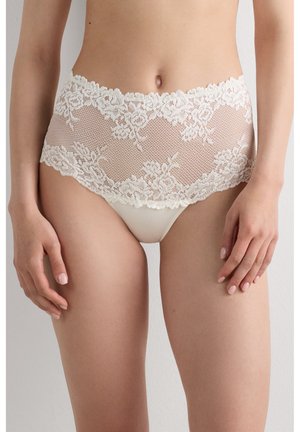 Žena nosící kalhotky s vysokým pasem z slonovinové krajky s květinovými vzory a síťovanými detaily, zobrazená od pasu po horní část stehen.