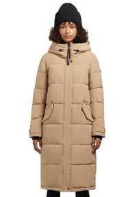 Béžová puffa bunda s horizontálnym prešívaním, veľká kapucňa, predný zips, dve bočné vrecká a čierna pletená čiapka na hlave modelky.