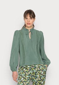 Blusa verde claro com colarinho franzido, abertura em forma de buraco da fechadura e detalhes em renda, combinada com calças escuras estampadas com flores. Textura suave e corte solto.