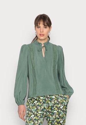 Blusa verde claro com colarinho franzido, abertura em forma de buraco da fechadura e detalhes em renda, combinada com calças escuras estampadas com flores. Textura suave e corte solto.