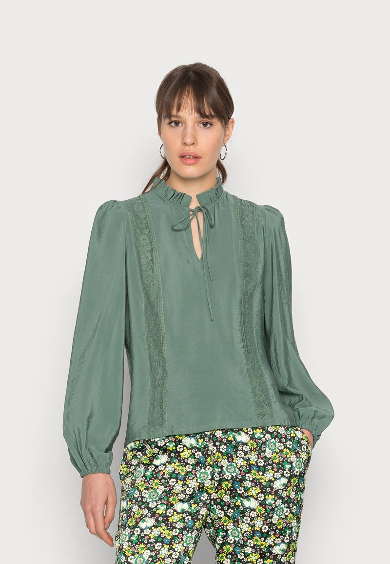 Blusa verde claro com colarinho franzido, abertura em forma de buraco da fechadura e detalhes em renda, combinada com calças escuras estampadas com flores. Textura suave e corte solto.
