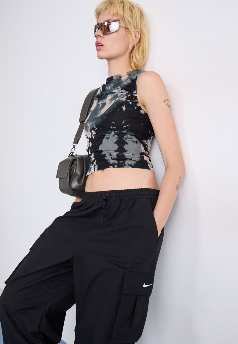 Top corto de tie-dye negro con cuello alto combinado con pantalones cargo negros que tienen bolsillos laterales y un logo de Nike en blanco. Los accesorios incluyen gafas de sol y un pequeño bolso de hombro.