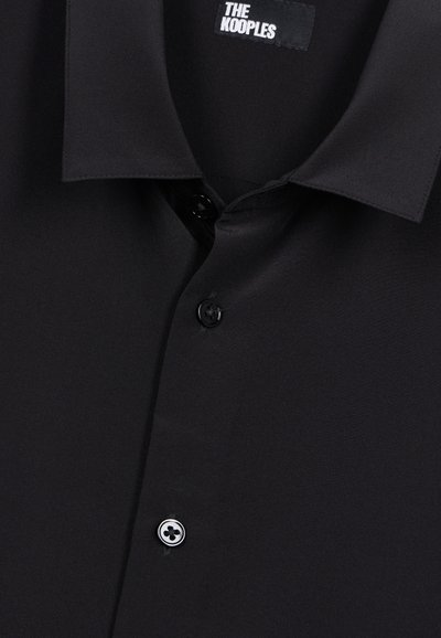 Sort skjorte med klassisk krave, knaplukning foran og en glat tekstur. Bemærkelsesværdige detaljer inkluderer et mærketikette og kontrasterende knapdesign.