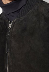 Giacca bomber in suede nera con colletto a coste, dotata di una cerniera argentata sul davanti e una texture liscia.
