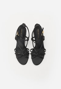 Sandales noires à lanières avec une finition en cuir lisse, semelle plate, accents noués, et une sangle de cheville ajustable dotée d'un fermoir de couleur dorée.