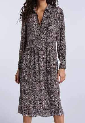 Femme portant une robe midi à manches longues, à col et boutonnée, avec un motif tacheté noir et beige.