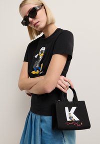 Schwarzes Grafik-T-Shirt mit einem Cartoon-Duck in einem Mantel, kombiniert mit einer kleinen schwarzen Handtasche, die ein großes weißes "K" und das Cartoon-Enten-Design zeigt.