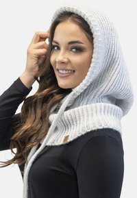 Căciulă tricotată de culoare gri deschis, cu o textură ribbată, având un guler lat și o etichetă din piele, așezată peste un top negru.