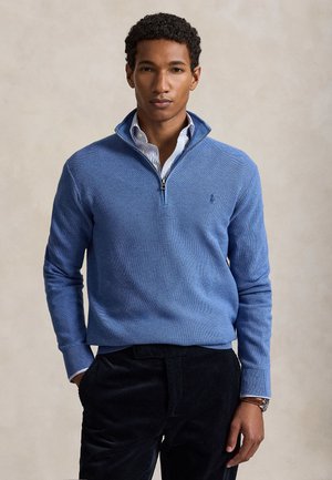 Jeune homme portant un pull bleu clair à fermeture éclair jusqu'à mi-poitrine sur une chemise rayée et un pantalon noir, debout avec une main dans la poche.