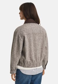 Woman wearing a gray speckled wool jacket with a dark brown collar, layered over a striped shirt and blue jeans, facing away.