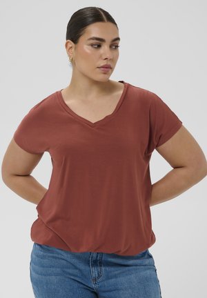 Kvinde med mørkt hår bundet tilbage, iført guldringe som øreringe, en rustfarvet V-hals trøje og blå jeans, stående med hænderne bag ryggen.