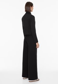Schwarzes maßgeschneidertes Outfit mit einer kurz geschnittenen Jacke und weiterbeinigen Hosen. Glatter Stoff mit tailliertem Design und klaren Linien. Schuhe mit einer leichten Sohle.