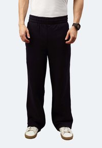 Schwarze Jogginghosen mit einem elastischen Bund, lockerer Passform und seitlichen Taschen, kombiniert mit weißen Sneakers. Glatte Textur ohne sichtbare Muster.