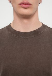 Καφέ βαμβακερό μπλουζάκι με στρογγυλή λαιμόκοψη, με μαλακή υφή και εμφανείς ραφές γύρω από το γιακά. Χωρίς σχέδια ή γραφικά.