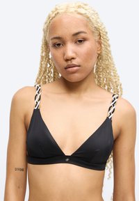 Γυναίκα με ξανθά πλεγμένα μαλλιά που φοράει μαύρο σουτιέν με μαύρα και λευκά πλεγμένα λουριά, εμφανές τατουάζ με τη λέξη "DIVINE" στο αριστερό της χέρι.