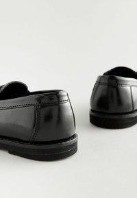 Zapatos de cuero negro con un acabado brillante, forma redondeada y suelas negras contrastantes. Se puede ver el detalle de la costura a lo largo de las costuras.