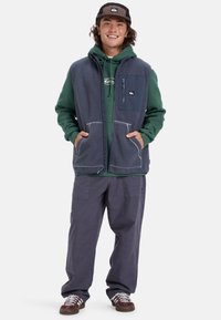 Fleeceväst i marinblått med en dragkedja på bröstfickan, gråa detaljer och två framfickor, lager på lager över en grön huvtröja och lösa grå byxor.