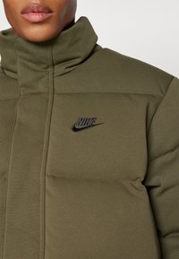 Giacca puffer Nike di colore verde oliva, con colletto alto, texture trapuntata e logo ricamato nero sul petto.