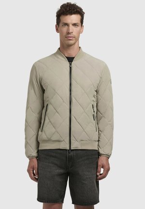 Gepolsterte beige Bomberjacke mit einem Frontreißverschluss, geripptem Kragen und seitlichen Reißverschlusstaschen; kombiniert mit dunklen Jeansshorts.