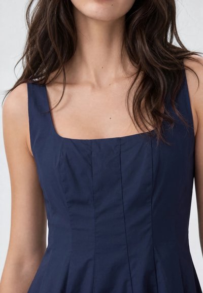 Femme portant une robe bleu marine sans manches avec un décolleté carré et des détails de coutures visibles sur le corsage.