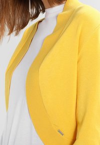 Gele gebreide cardigan met een open ontwerp en geschulpte rand, gedragen over een wit topje. Voorzien van een klein zilverkleurig metalen detail. Zachte textuur.