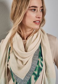 Sciarpa crema con motivo geometrico verde e blu lungo i bordi. Realizzata in tessuto leggero con una forma drappeggiata e rilassata.