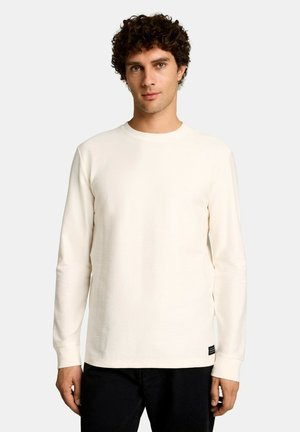 Jeune homme aux cheveux foncés et bouclés, portant une chemise unie à manches longues couleur écru et un pantalon noir, debout devant un fond uni.