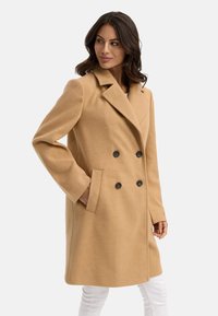 Beige dubbelknäppt kappa med notchkrage, svarta knappar och sidofickor, tillverkad av textil med struktur, med en skräddarsydd passform.