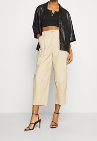 Chemise courte en similicuir noir superposée à un crop top noir et un pantalon large beige, complétée par des sandales à talons hauts noires à fines lanières.