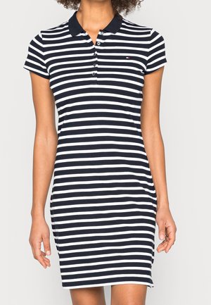 Frau trägt ein schwarz-weiß horizontal gestreiftes Polo-Kleid mit kurzen Ärmeln, Knöpfen und einem kleinen Logo auf der Brust.