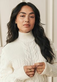 Chunky cream-colored cable knit sweater with a high turtleneck and ribbed texture, featuring long sleeves and oversized cuffs.