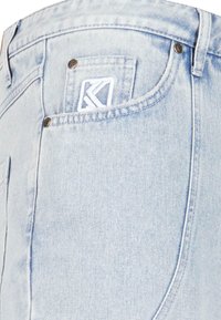 Pantalones vaqueros de mezclilla azul claro con un bolsillo trasero que presenta un logo bordado en blanco y remaches metálicos, mostrando un ajuste y textura clásicos.
