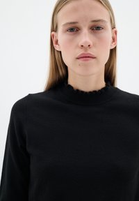 Svart stickad tröja med hög hals och volangkant. Texturen är mjuk och designen är enkel och figurnära.