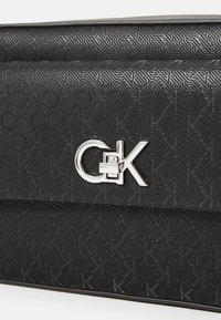 Borsa nera in tessuto con un motivo in rilievo e un logo argentato. Il design è elegante con una forma rettangolare e una finitura liscia.