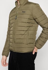 Olivgrön vadderad jacka med hög krage, dragkedja fram och sidfickor. Har sydda detaljer och en FILA-logga på bröstet.