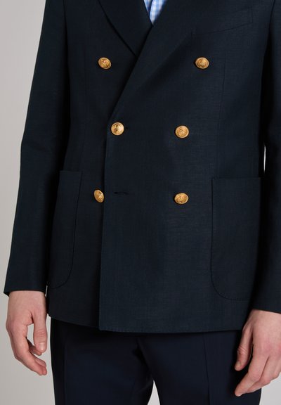 Mann trägt marineblauen doppelt geknöpften Blazer mit sechs goldenen Knöpfen und aufgesetzten Taschen vorne, kombiniert mit dunkler Hose und hellblau kariertem Hemd.