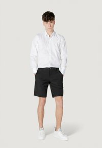 Camisa blanca de botones, pantalones cortos negros y zapatillas blancas. Los pantalones cortos son ajustados, con bolsillos y cierre de botón. Mangas cortas en la camisa.