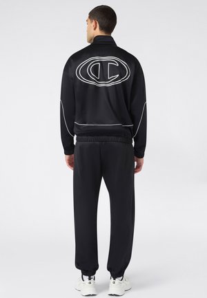 Hombre de espaldas vistiendo un chándal negro con logo blanco y detalles con ribetes, y zapatillas blancas con logo negro.