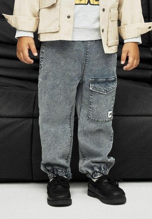 Pantalons cargo en denim bleu délavé avec une taille élastiquée. Présente une grande poche latérale et une texture usée, associés à des chaussures noires à lacets.