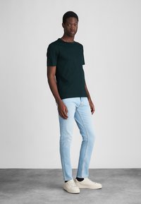 Tmavo zelené bavlnené tričko kombinované so svetlomodrými džínsy a béžovými teniskami. Tričko má okrúhly výstrih a krátke rukávy.