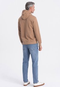 Felpa marrone con cappuccio regolabile, abbinata a jeans di denim blu chiaro e sneaker bianche. Texture liscia, vestibilità casual e design semplice.