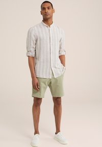 Svetlo béžová pruhovaná košeľa s gombíkmi a stojacím golierom, v kombinácii so zelenými šortkami a bielymi teniskami. Neformálny outfit s voľným strihom.