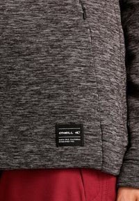 Szary, teksturowany sweter z kieszenią na zamek, z czarną metką marki na dole. Materiał ma melanżowy wzór.