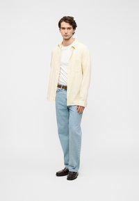 Ανοιχτό κίτρινο πουκάμισο με κουμπιά, λευκή μπλούζα, ανοιχτό μπλε τζιν και σκούρα καφέ παπούτσια. Απλός σχεδιασμός με μια τσέπη στο πουκάμισο.